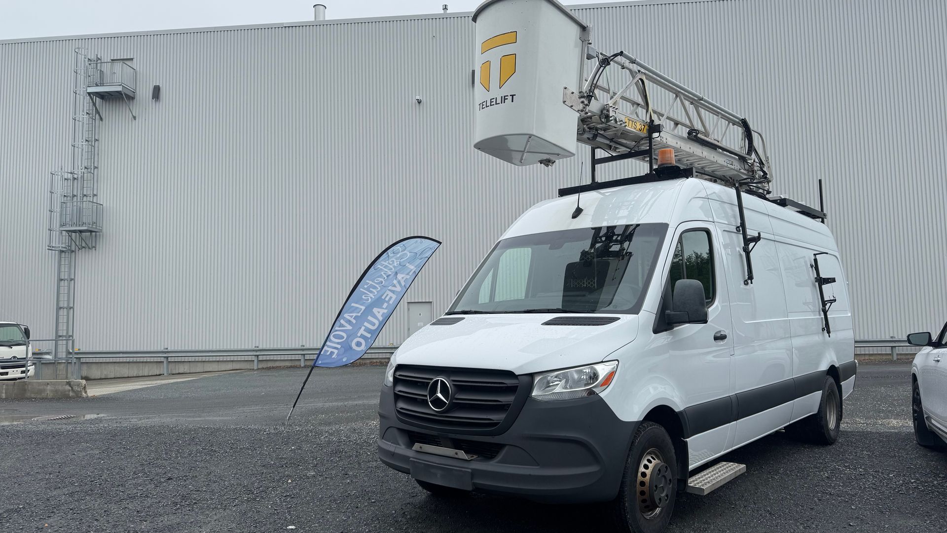 Fourgonnette blanche avec nacelle élévatrice garée devant un bâtiment blanc.