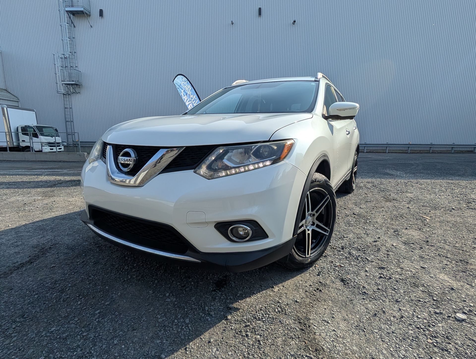 SUV Nissan Rogue blanc garé en extérieur. Jantes noires, portière ouverte, devant un grand bâtiment.