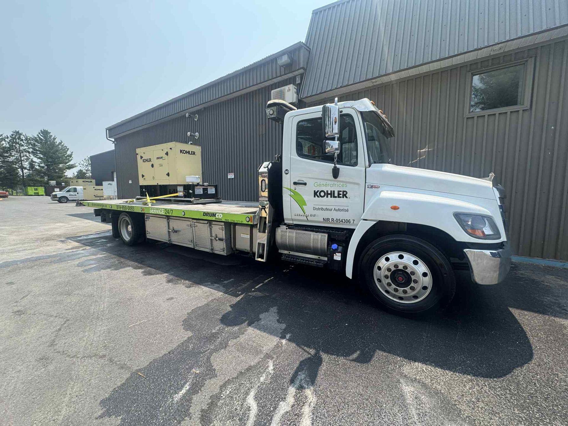 Camion à plateau blanc avec un générateur sur son plateau garé devant un bâtiment par une journée ensoleillée.
