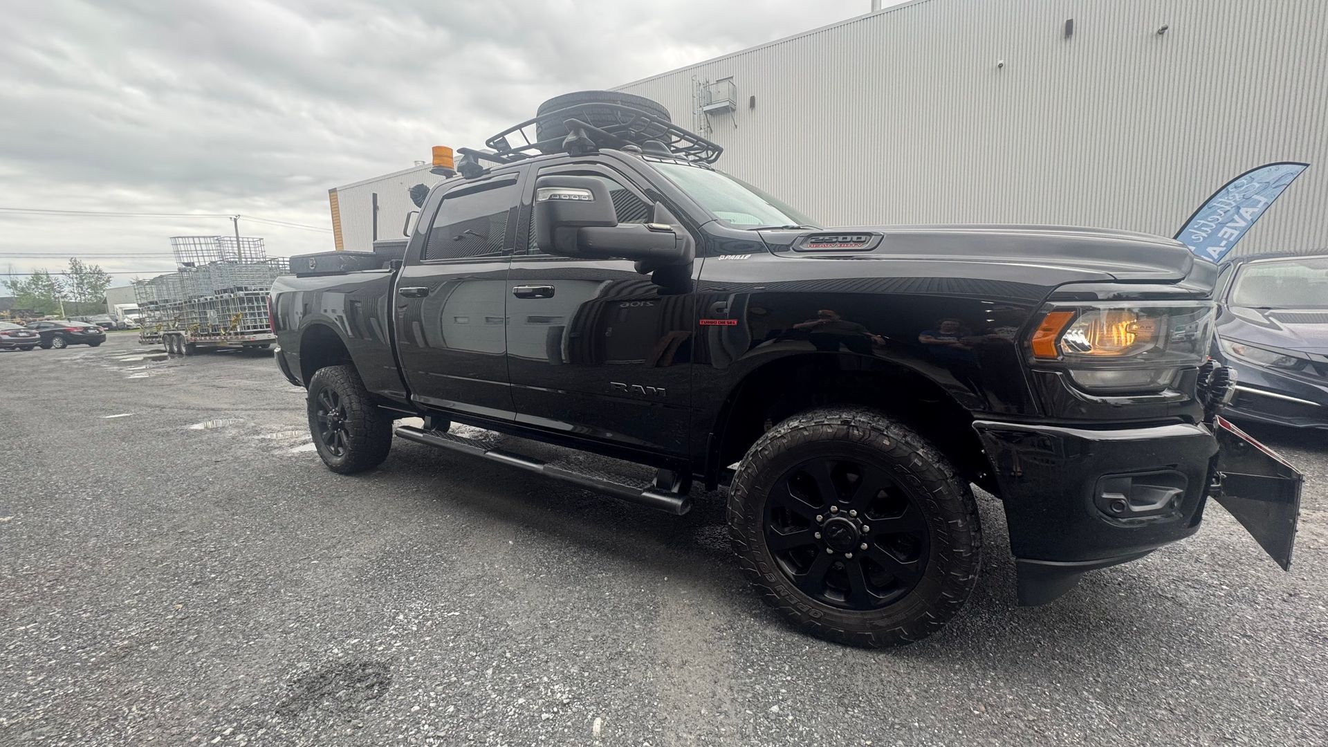 Pick-up noir avec chasse-neige, garé sur l'asphalte. Une galerie de toit permet de ranger du matériel. Journée nuageuse.