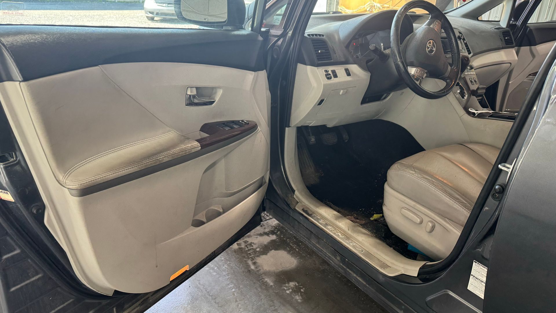 Intérieur porte de voiture ouverte, sièges en cuir beige, tableau de bord noir, volant visible.