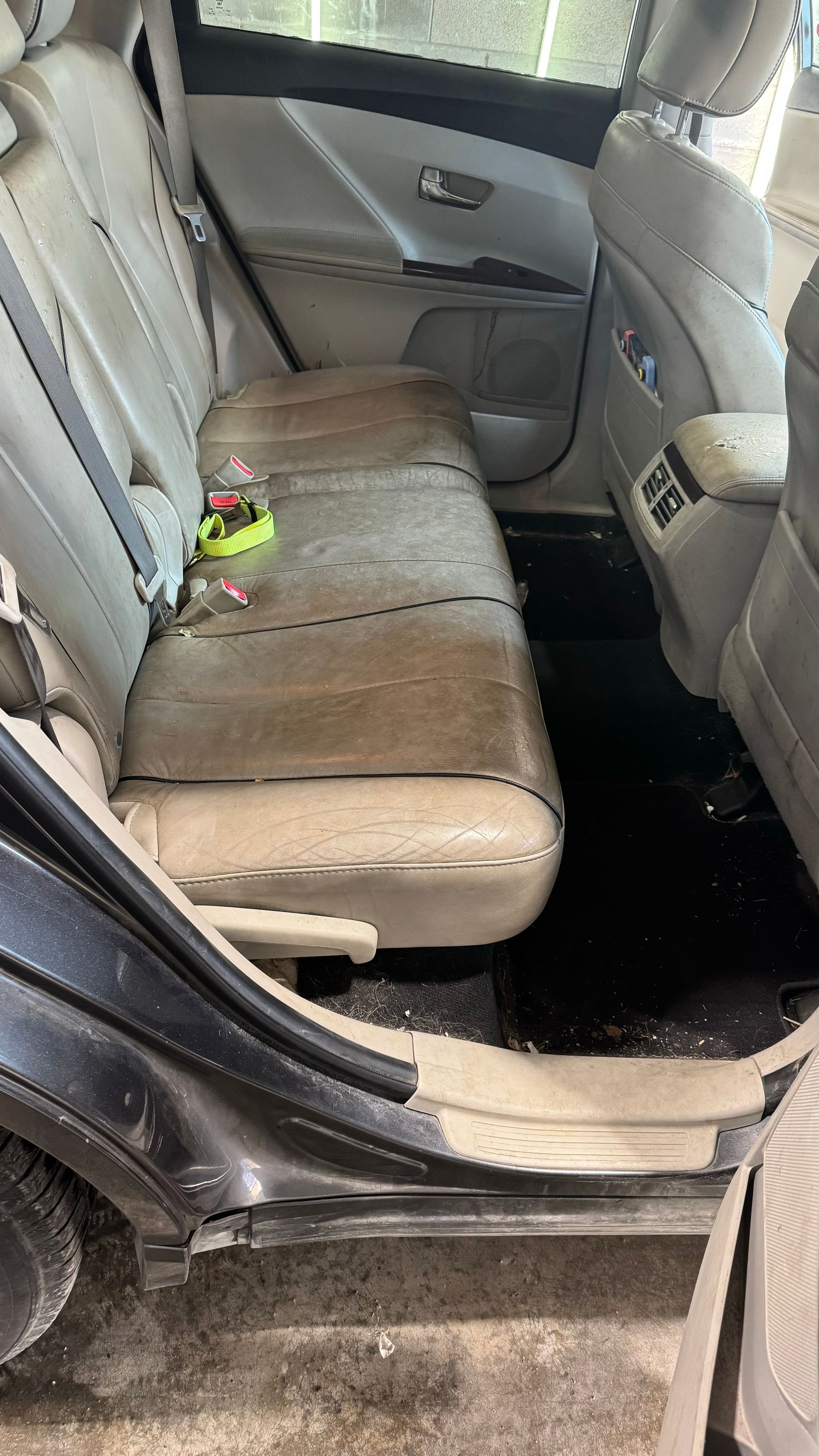La banquette arrière d'une voiture est sale. Sièges en cuir beige, tapis de sol noirs, cadre de porte visible.
