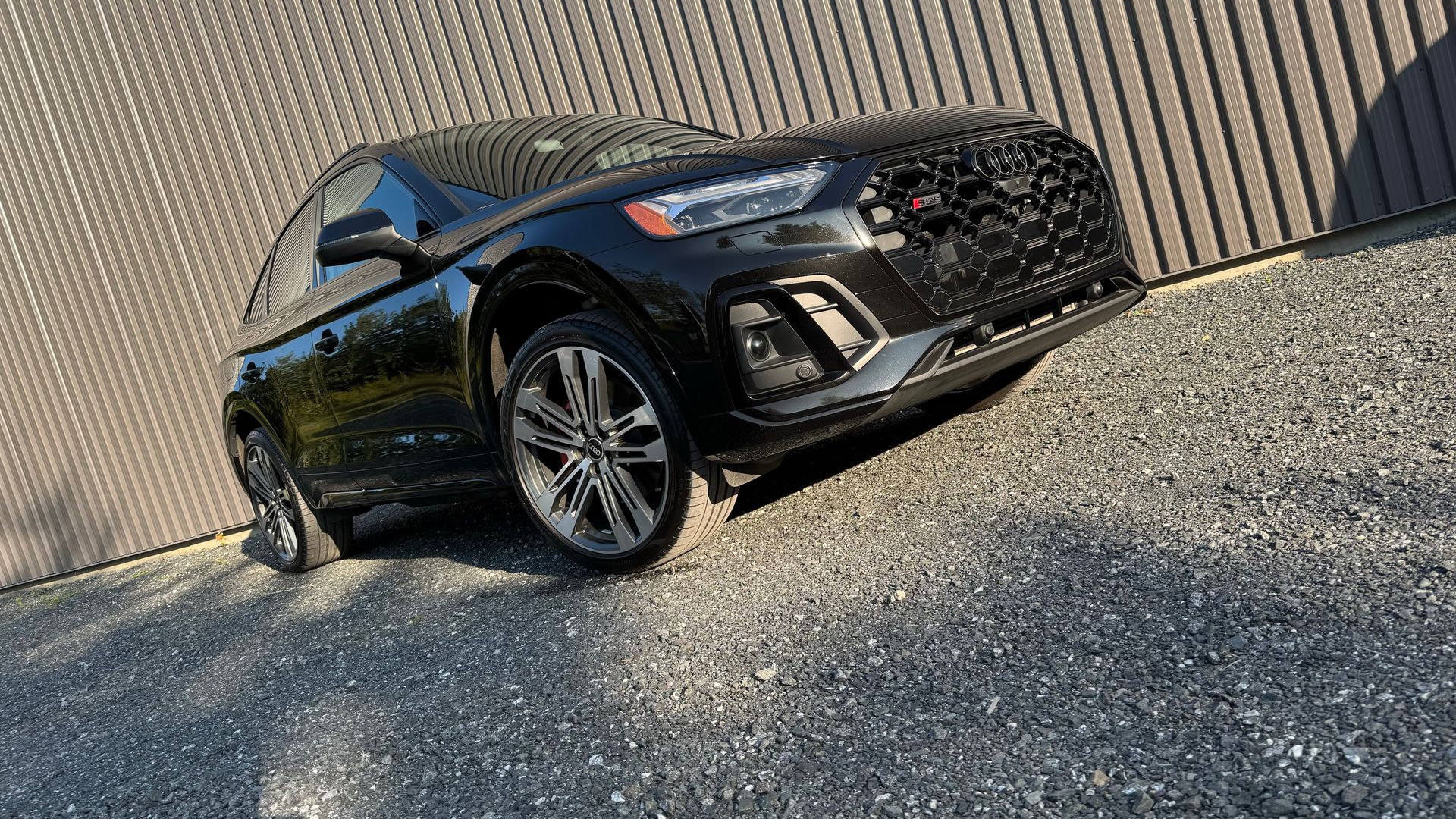 SUV Audi noir garé sur une surface de gravier à côté d'un mur en tôle ondulée.