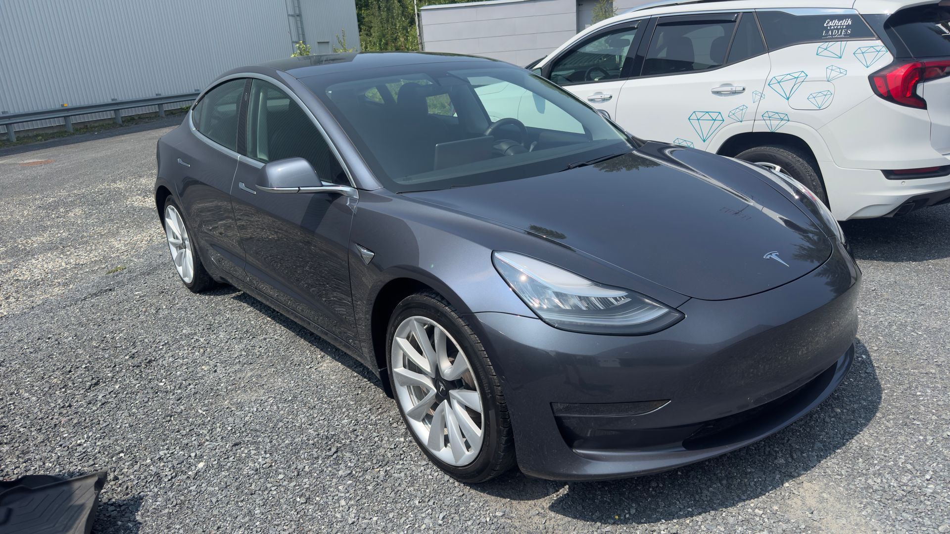 Berline Tesla Model 3 grise garée à l'extérieur à côté d'un SUV blanc sur du gravier.