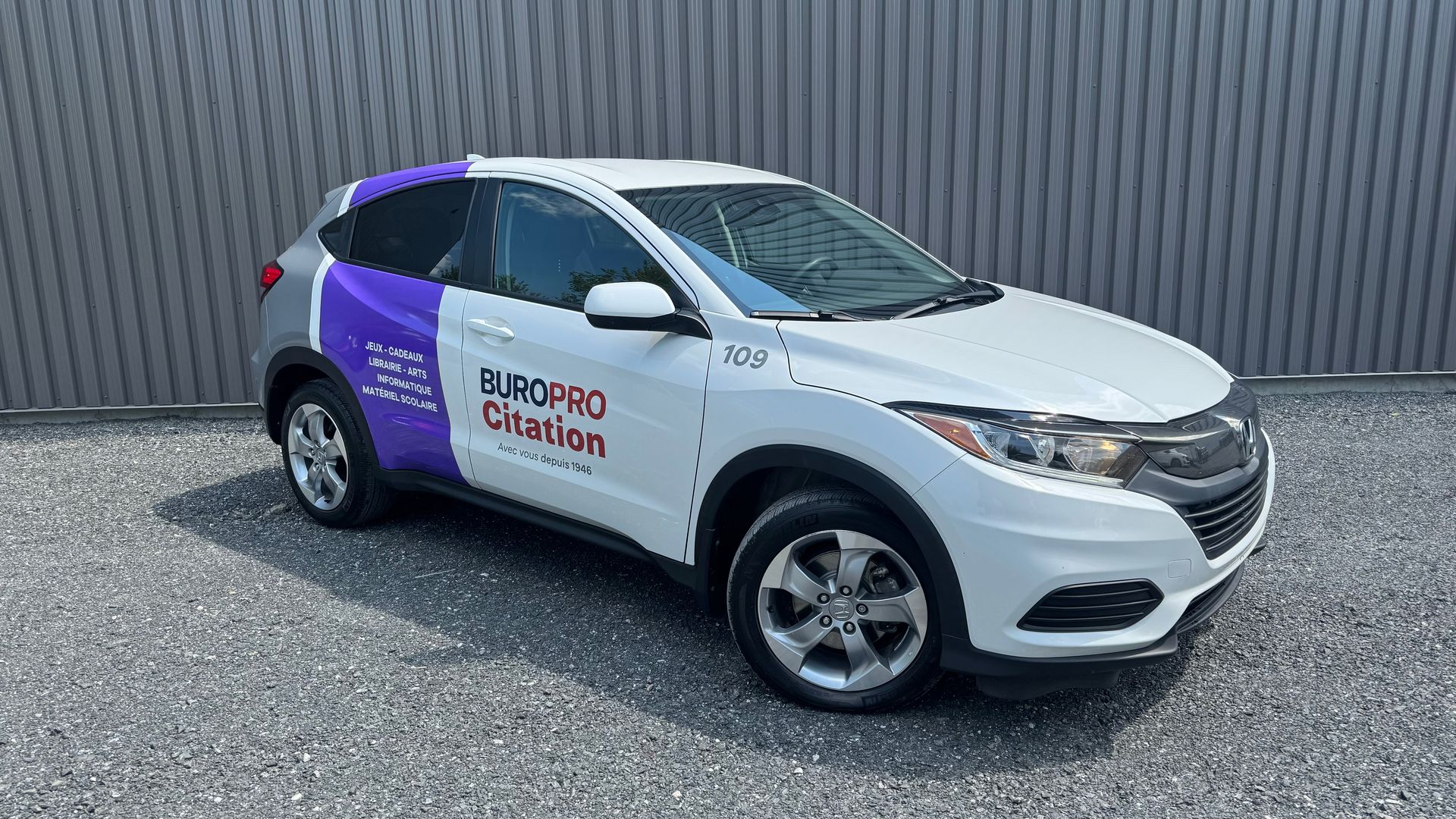 Honda HR-V blanc avec un habillage publicitaire violet et blanc garé devant un mur gris.