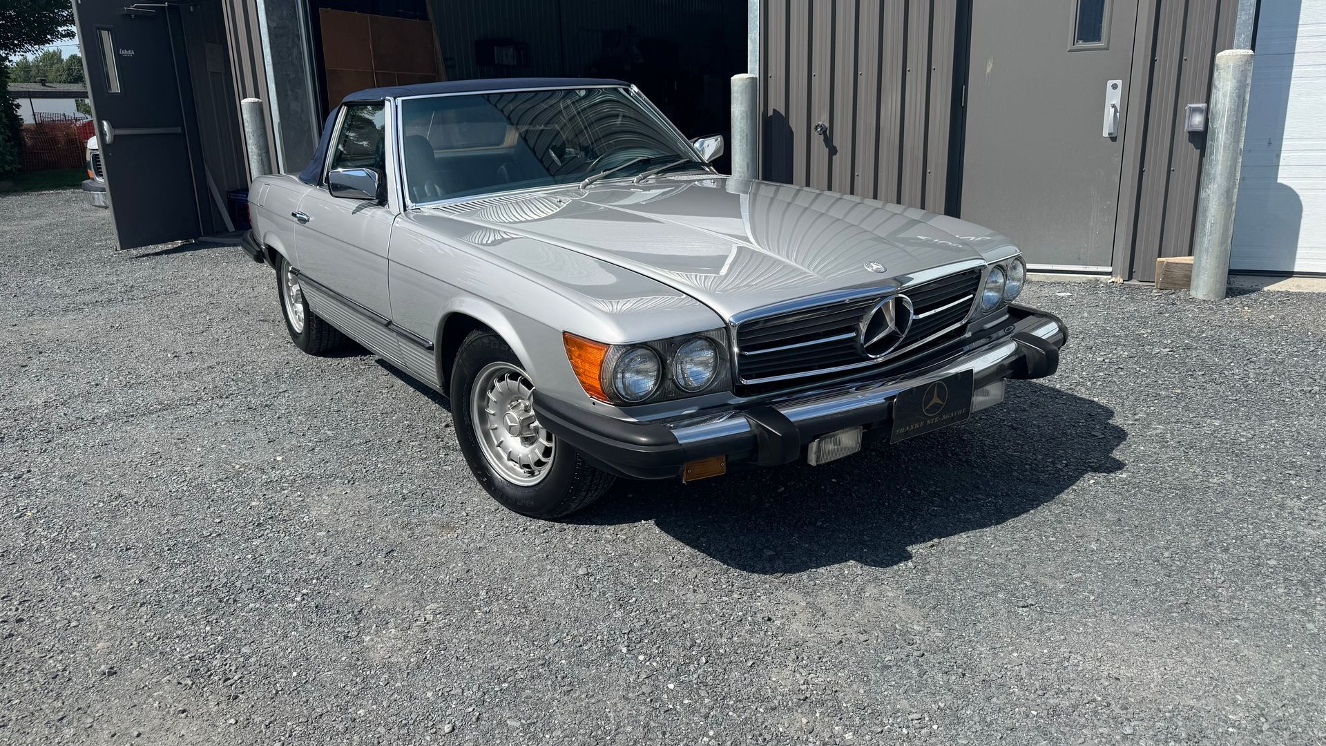 Mercedes-Benz SL cabriolet argentée garée sur du gravier, avec un toit sombre et un garage ouvert en arrière-plan.