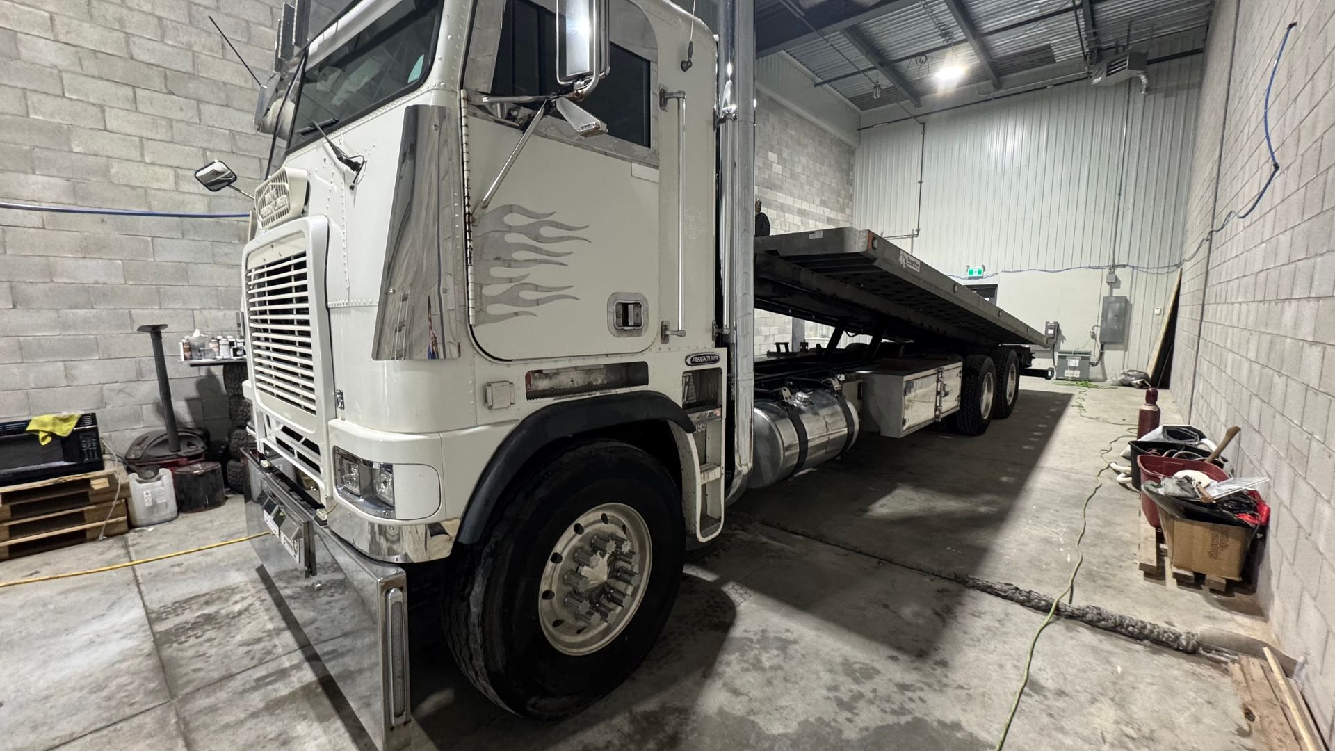 Semi-remorque blanc garé dans un garage. On y voit des détails chromés, un long plateau et des murs en briques.