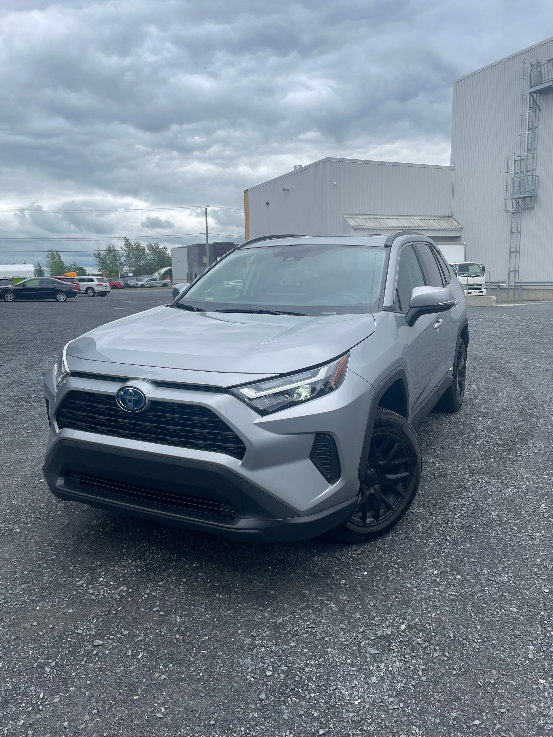 SUV hybride Toyota RAV4 argenté garé sur du gravier devant un bâtiment, ciel couvert.