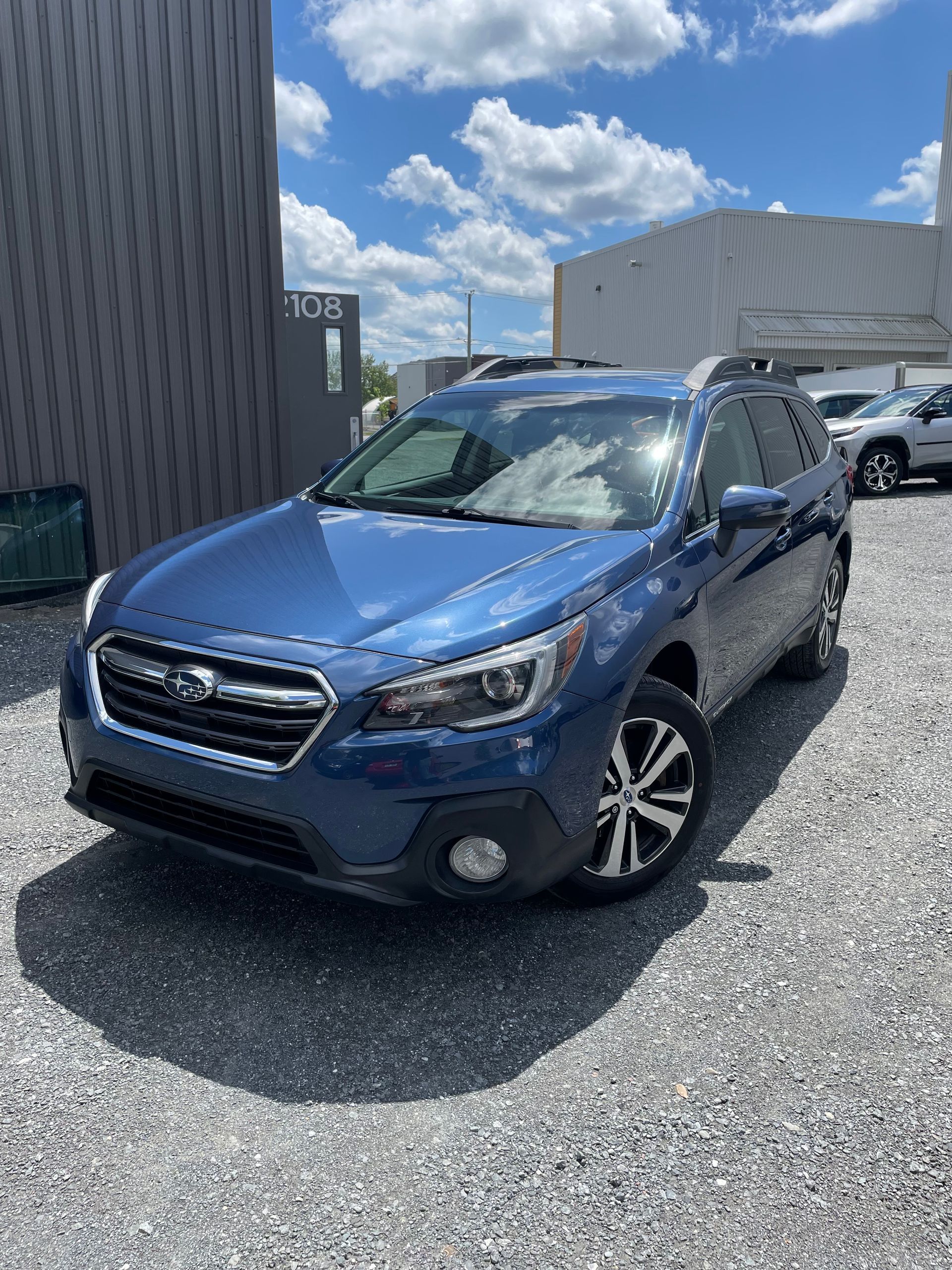 Subaru Outback bleu garé sur un terrain en gravier sous un ciel partiellement nuageux.
