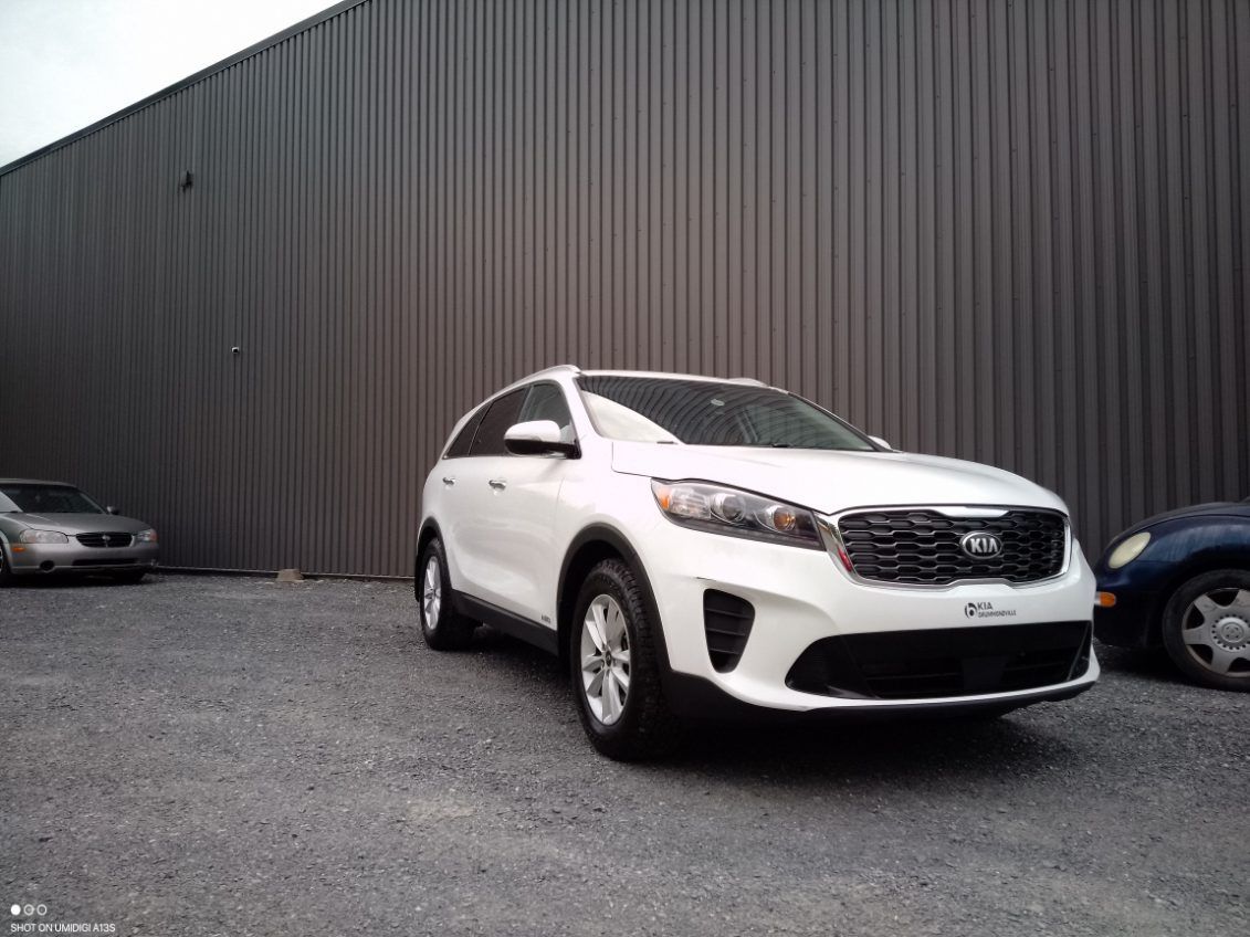 Un SUV Kia blanc garé devant un bâtiment gris ; une autre voiture à gauche et une bleue à droite.