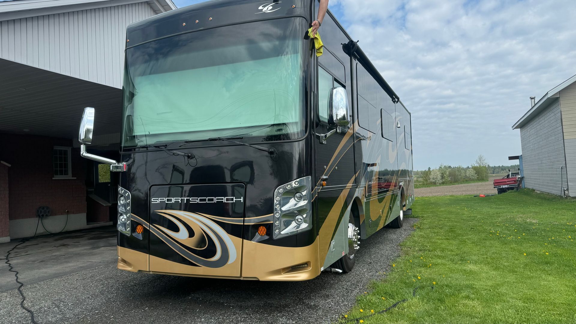 Camping-car noir et or garé dans une allée, à côté de l'herbe et des bâtiments, sous un ciel nuageux.
