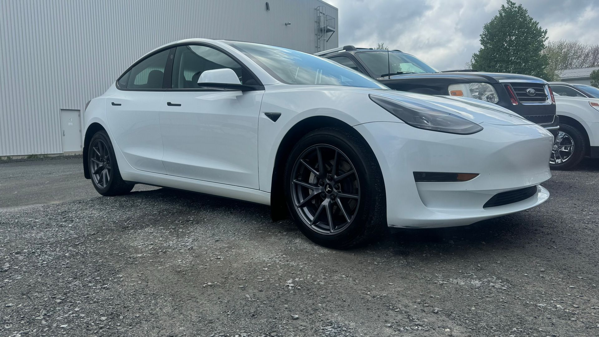 Tesla Model 3 blanche garée sur du gravier, avec des roues foncées. Une autre voiture est partiellement visible en arrière-plan.
