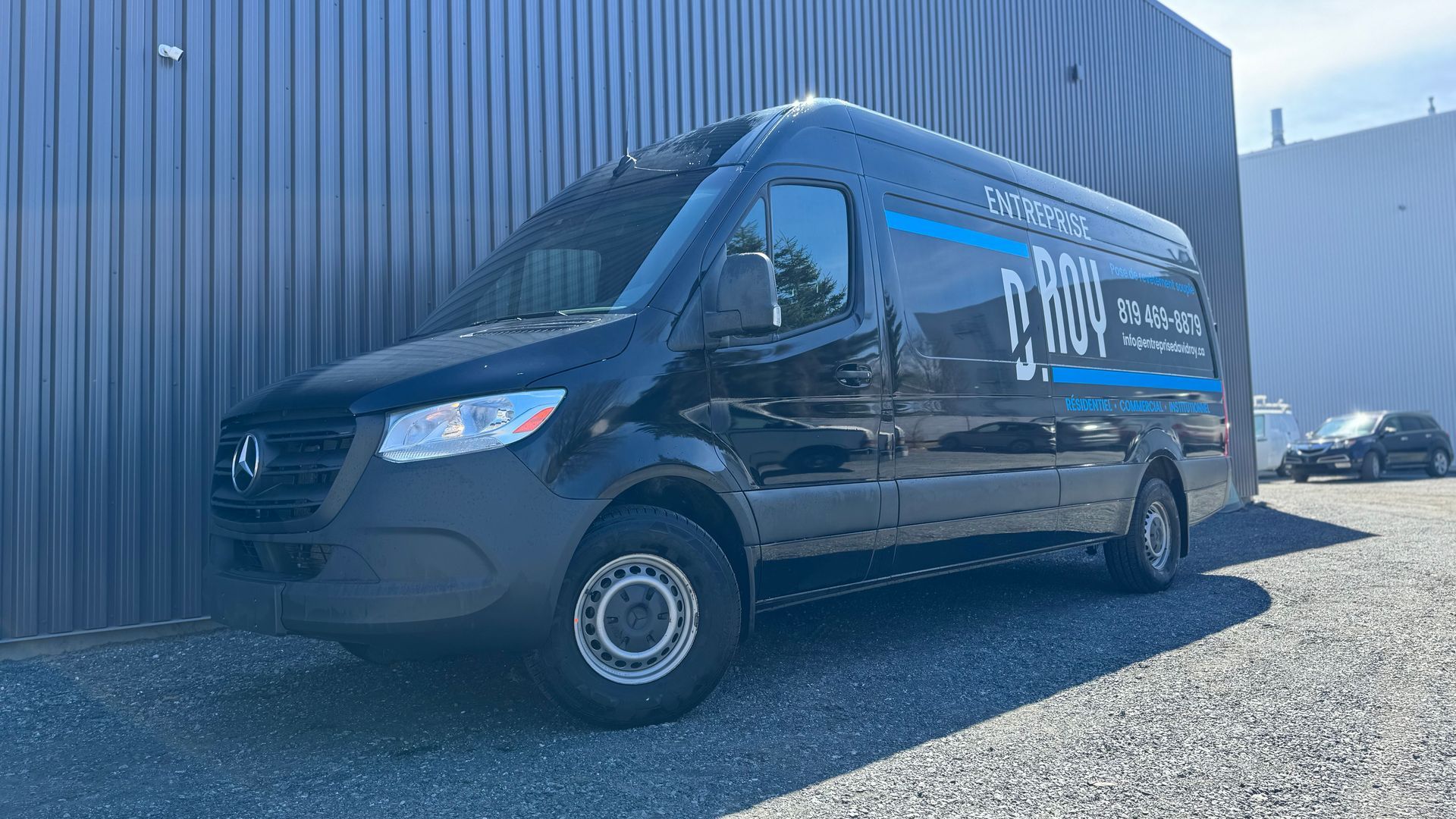 Fourgonnette Mercedes-Benz noire garée à côté d'un bâtiment gris. Elle arbore un lettrage et un logo bleus.