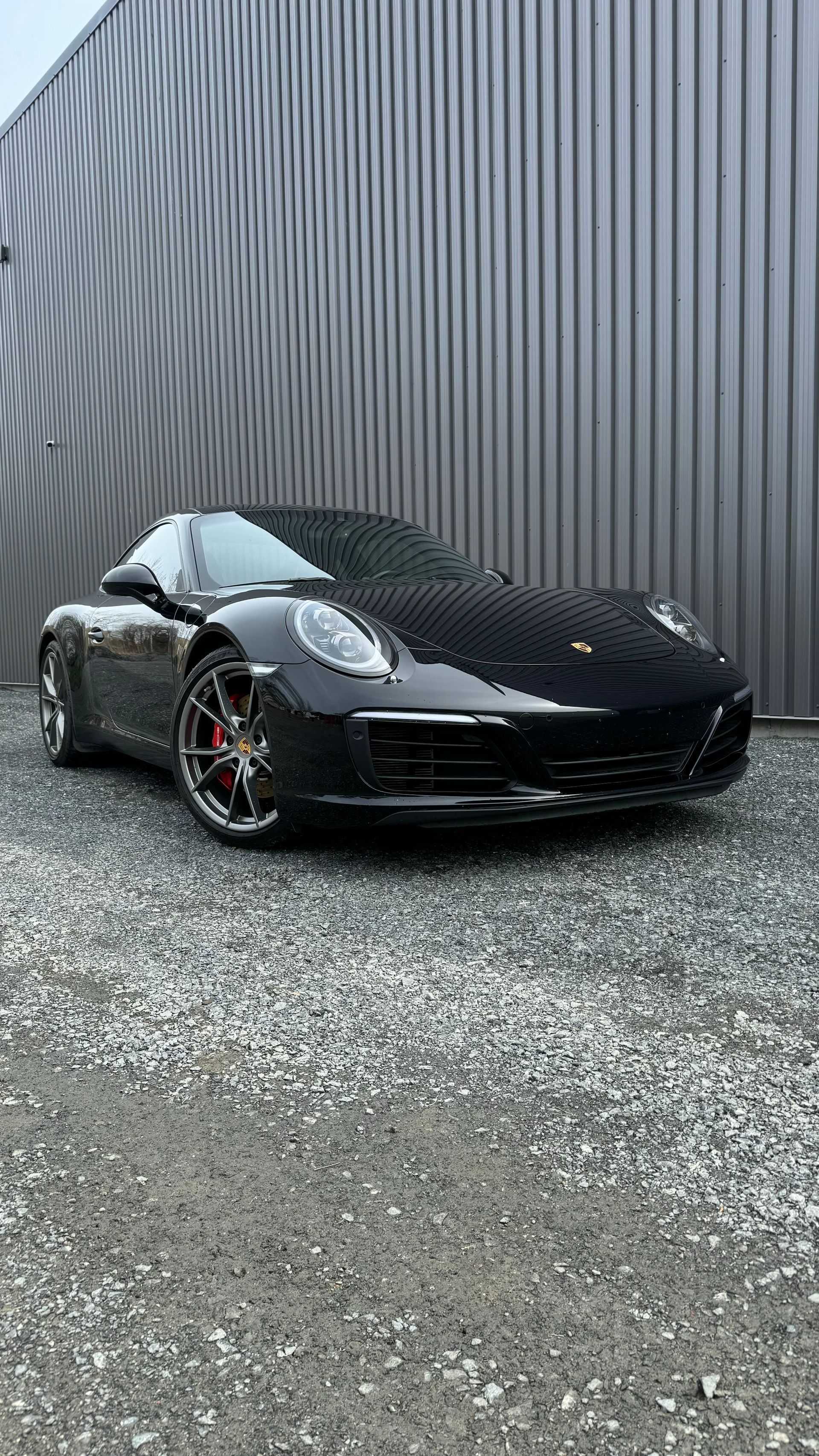 Porsche décapotable noire garée devant un mur en tôle ondulée sur une surface en gravier.