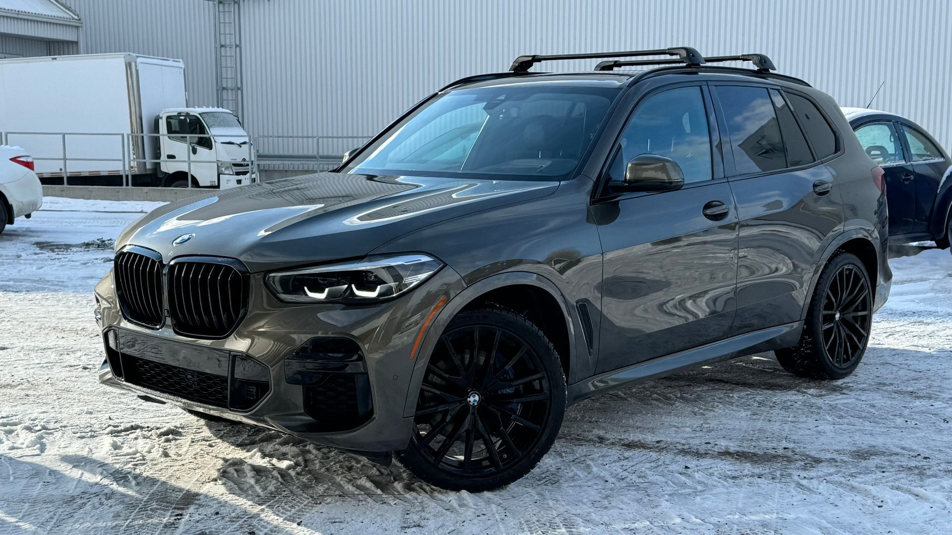 SUV BMW X5 gris foncé avec jantes noires et galerie de toit garée dans un terrain enneigé.