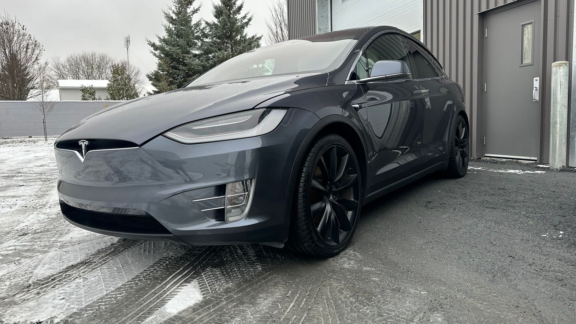 Tesla Model X gris foncé garée devant un bâtiment avec de la neige au sol.