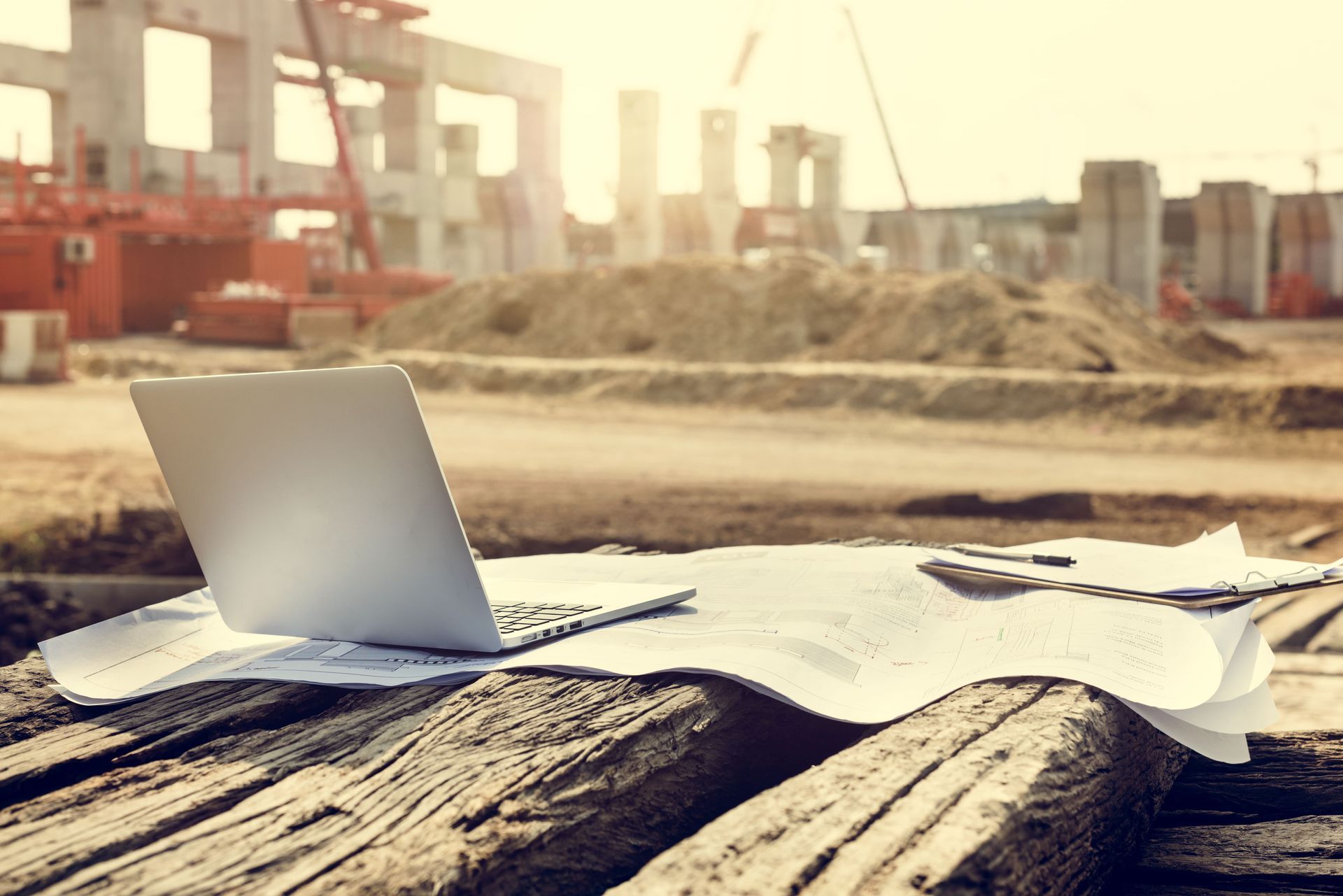 Laptop and blueprints on wooden beams at a construction site, bathed in sunlight.