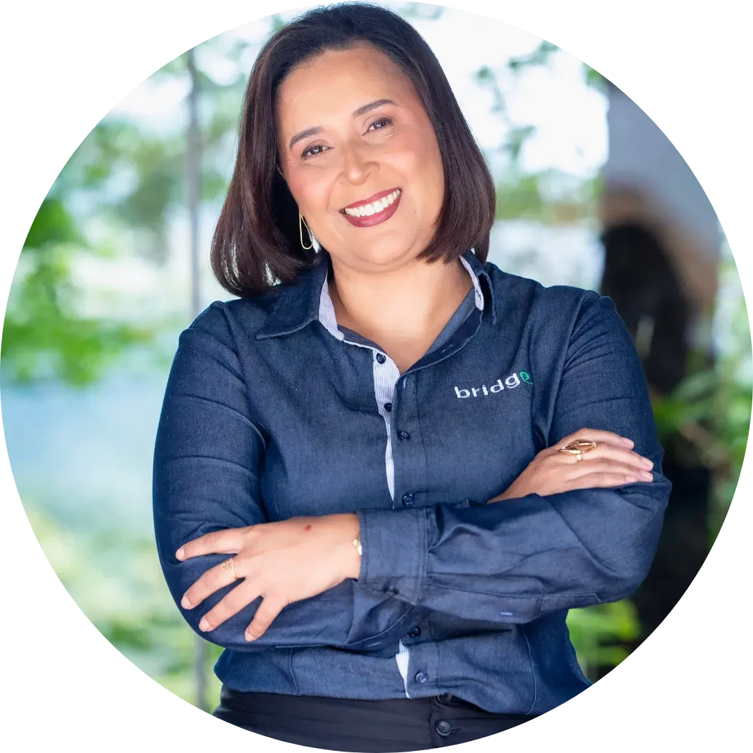 Uma mulher de camisa azul uniforme Bridge Gestão Social está sorrindo com os braços cruzados