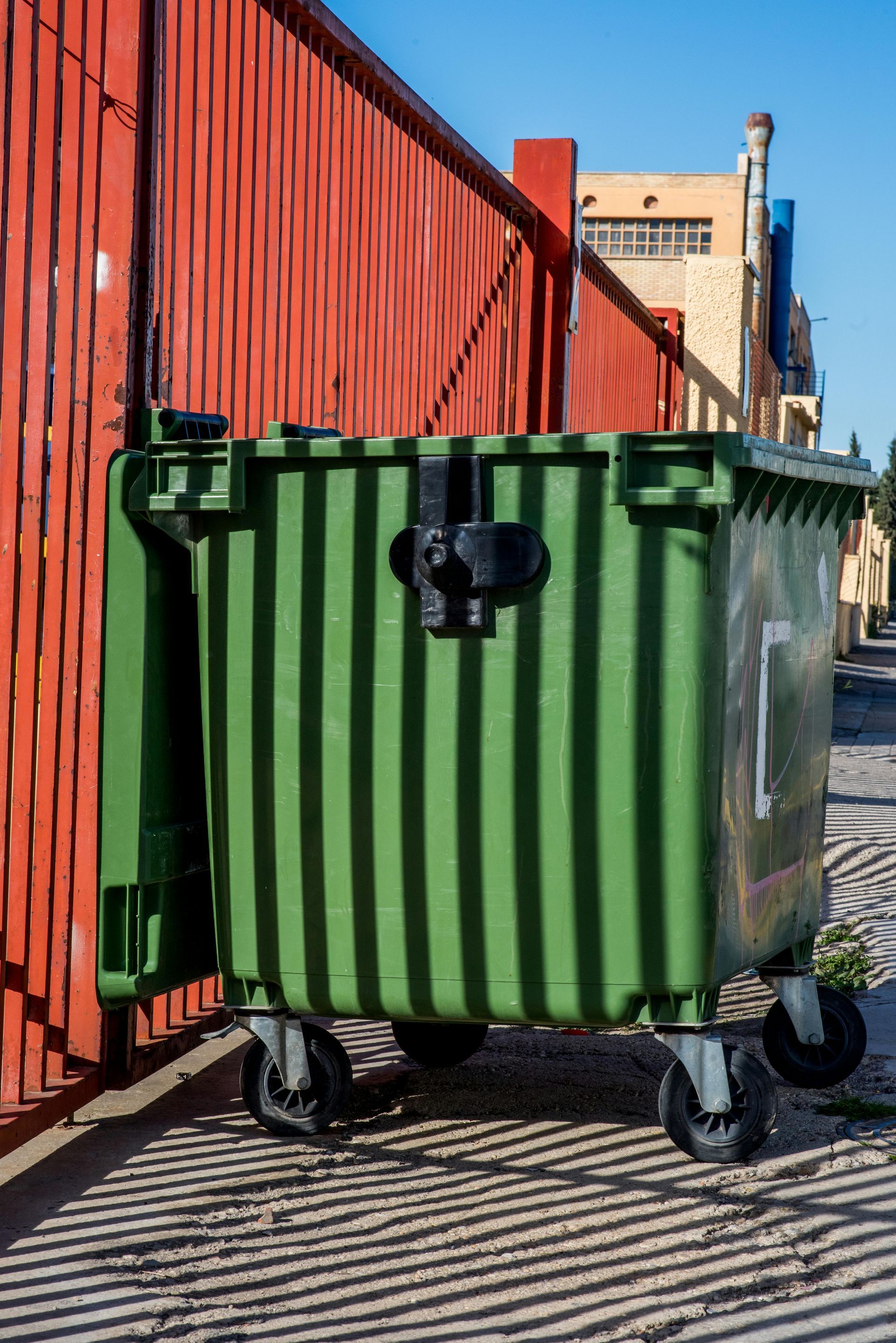 A green garbage can is parked in front of a red fence.