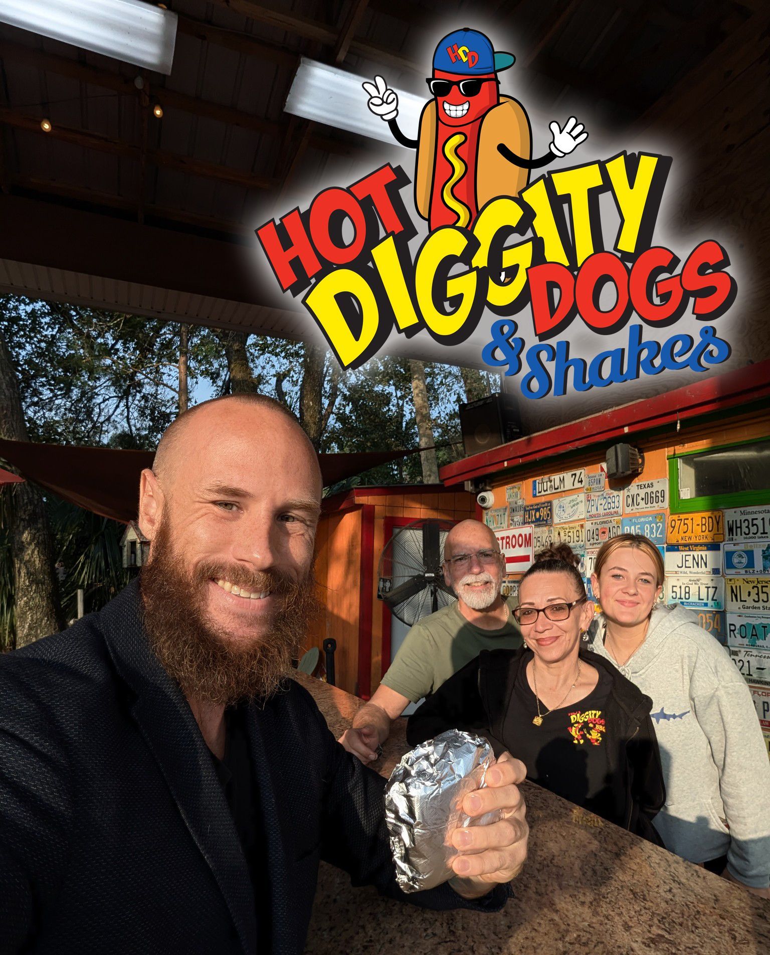 A man is holding a hot dog in front of a sign that says hot diggity dogs & shakes