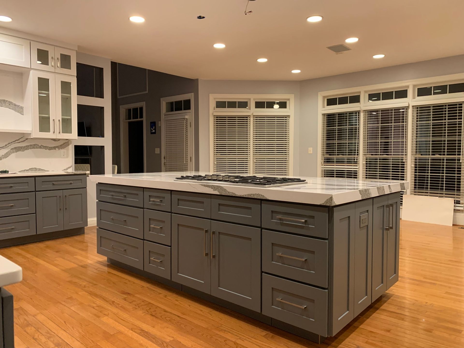 A gray kitchen island with a marble countertop. Gray cabinets surround it. Hardwood floors and white cabinets on the walls.