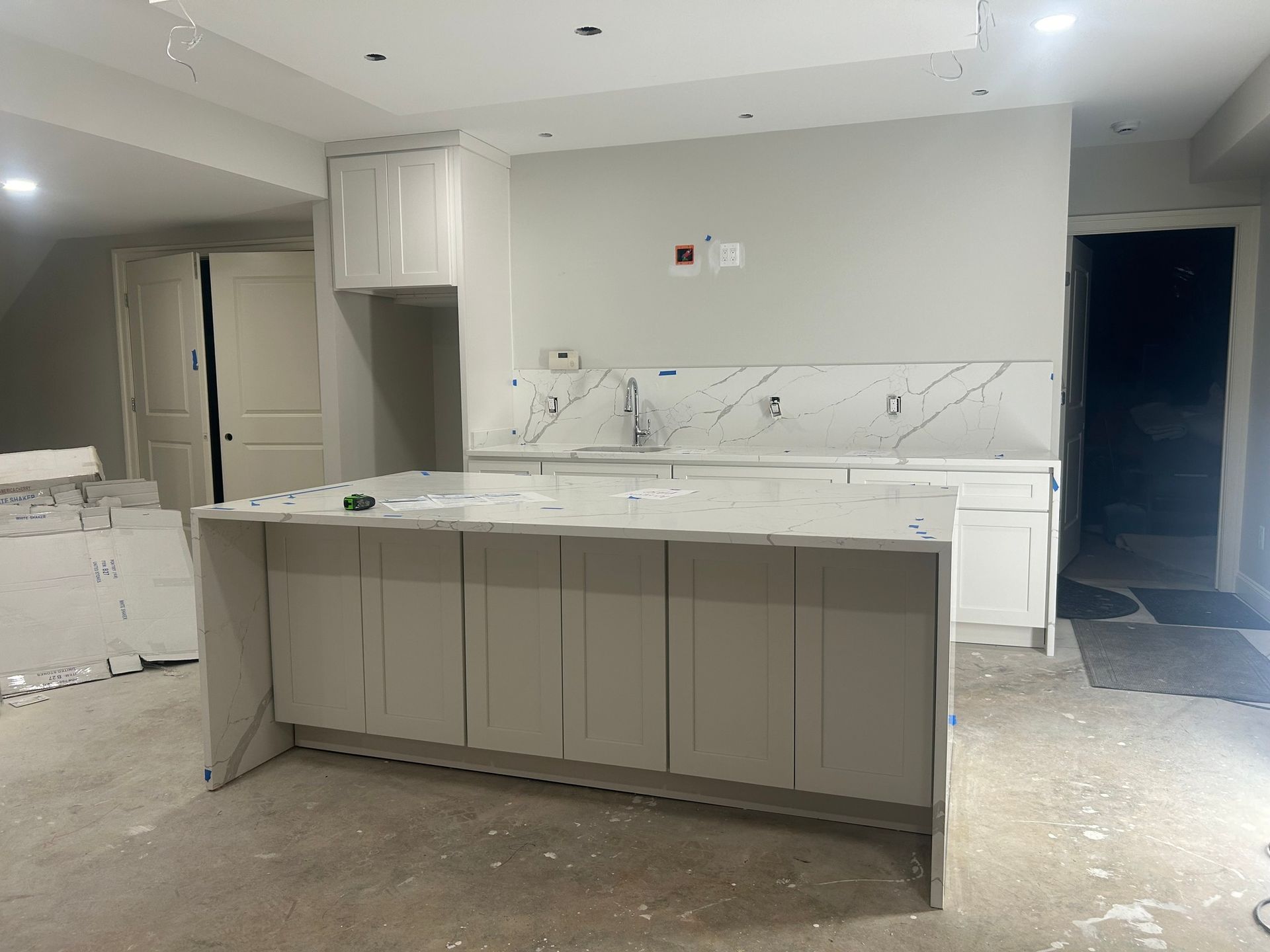 Kitchen under construction with white cabinets, island, and countertop. Walls are light gray; floor is unfinished concrete.