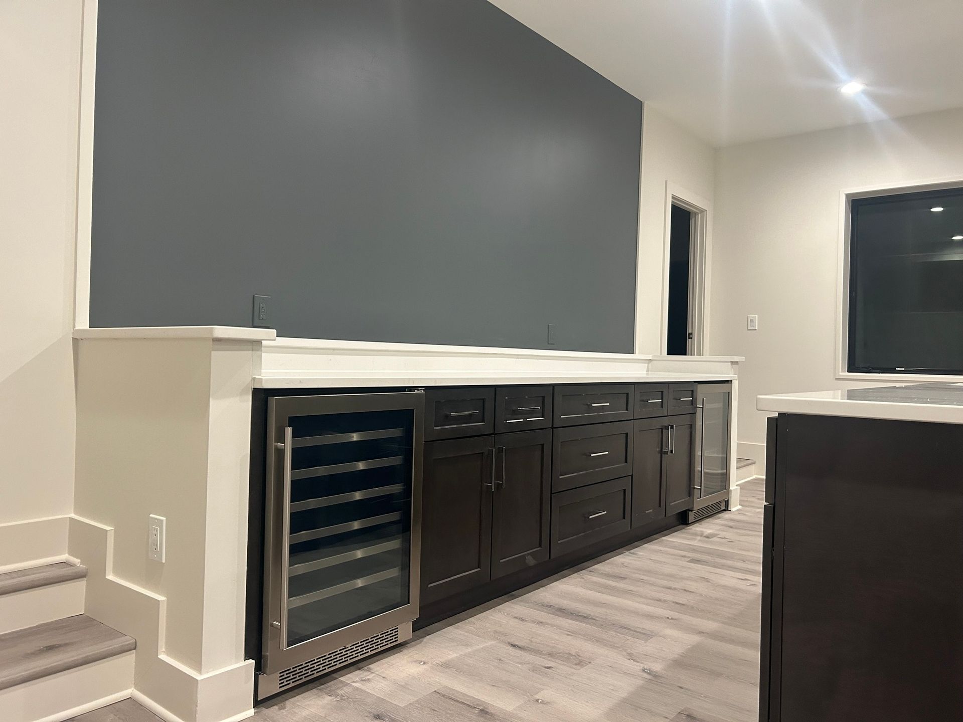 A built-in bar area with a wine fridge, dark cabinets, and a gray accent wall, set in a room with light flooring.