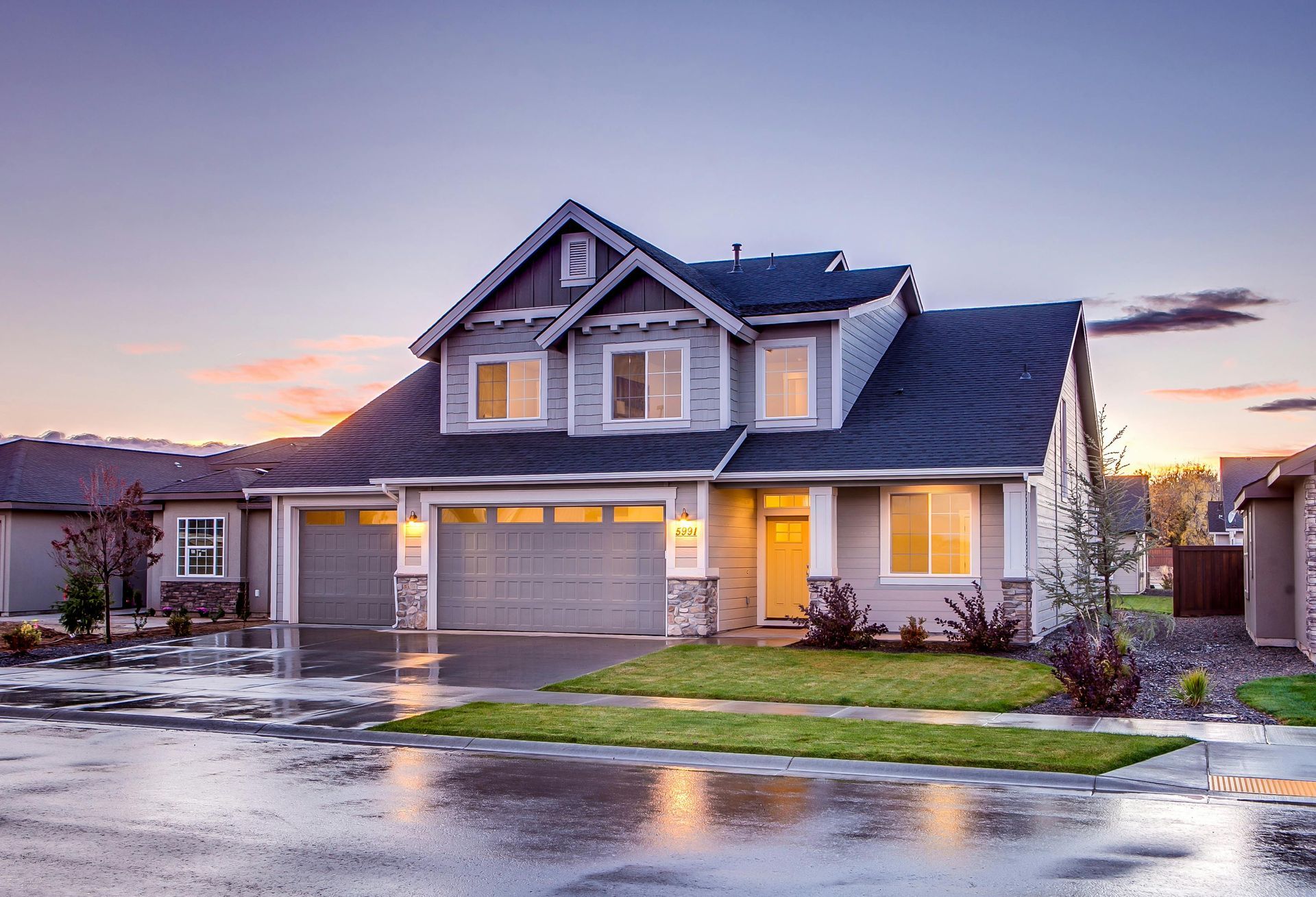Two-story gray house with a three-car garage at dusk; wet pavement reflects the sky.