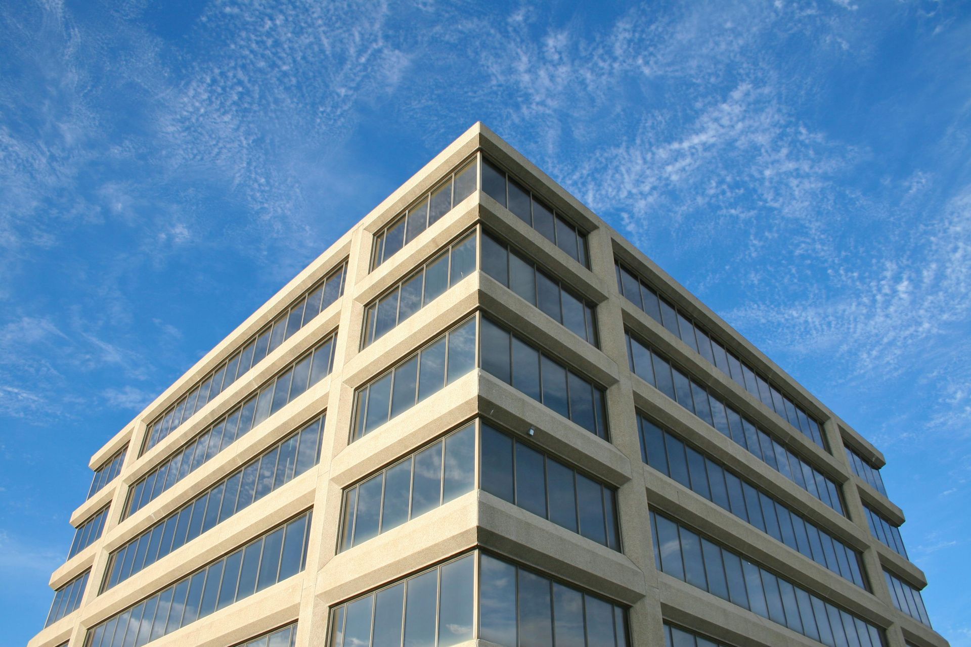 Modern office building, beige facade, glass windows, against a bright blue sky with wispy clouds.