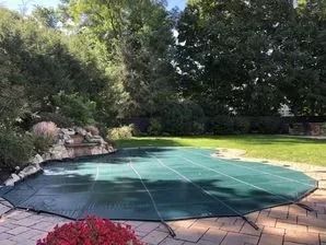 A green pool cover is sitting on top of a brick patio next to a swimming pool.