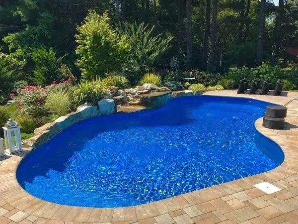 A large blue swimming pool is surrounded by trees and a brick patio.