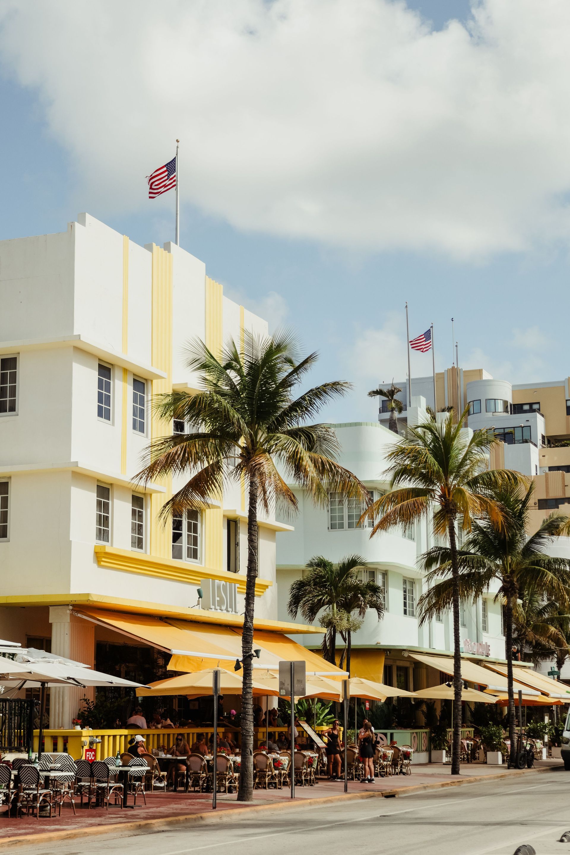 Palm-lined street with yellow-and-white buildings, outdoor café seating, and flags flying in a sunny seaside area