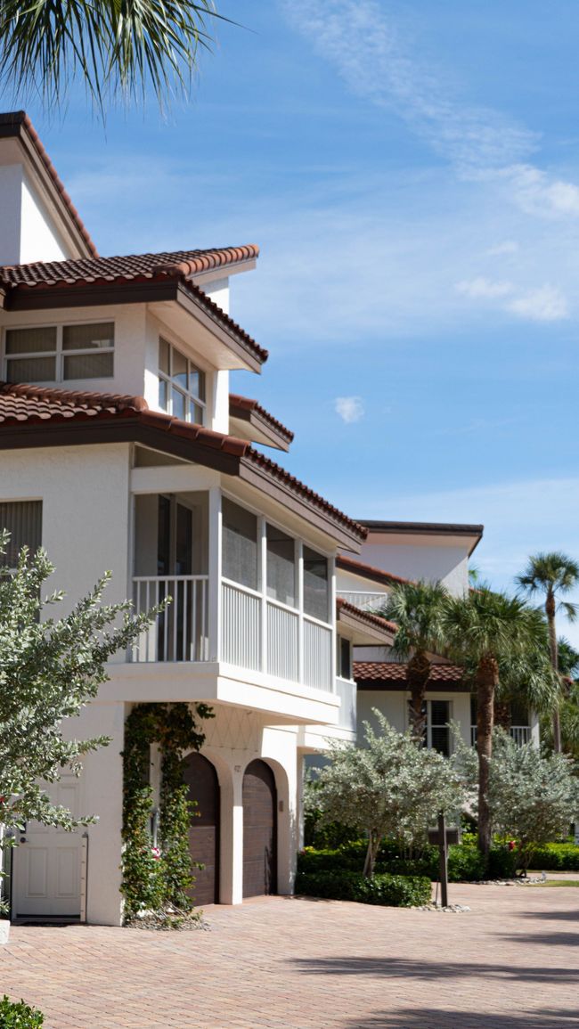 Mediterranean-style villa with white balconies and red tile roofs beside a gravel path under a blue sky