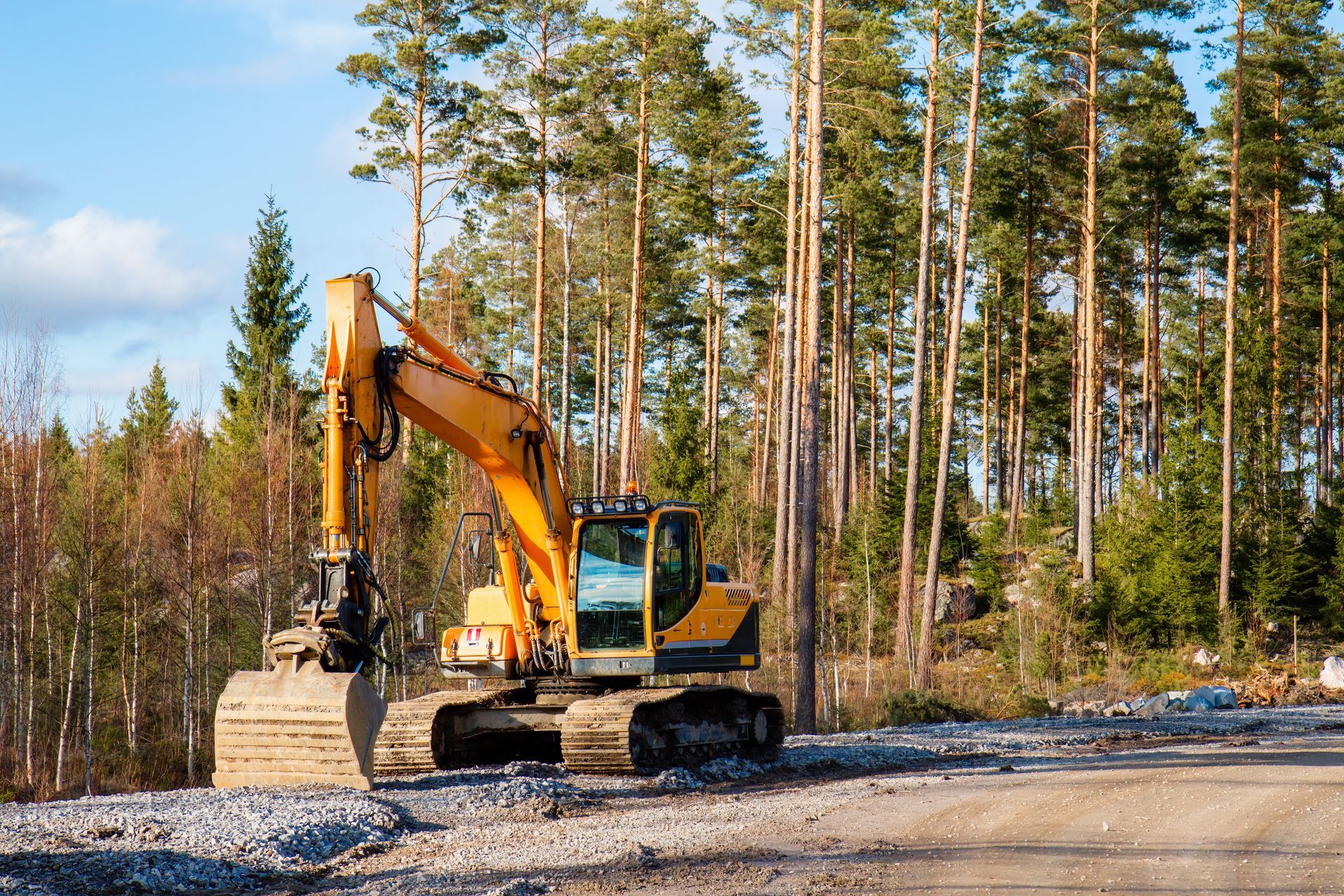 Une excavatrice jaune roule sur un chemin de terre au milieu d'une forêt.