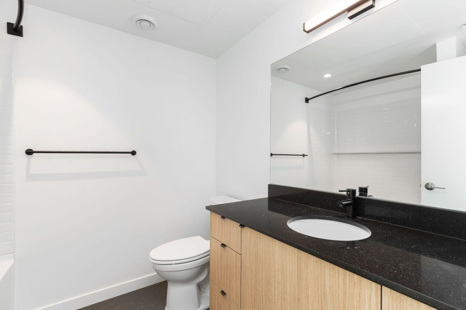 Modern bathroom with white walls, black countertop, light wood vanity, and a large mirror.