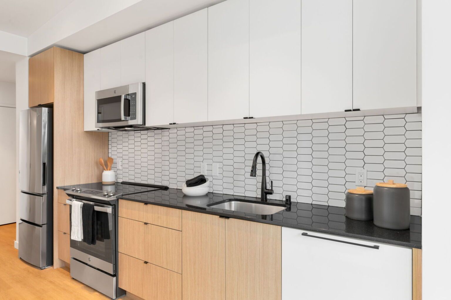 Modern kitchen with white and wood cabinets, black countertops, and a stainless steel fridge.
