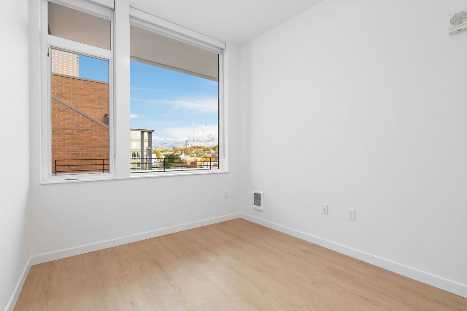 Empty, bright room with a large window overlooking cityscape. Light wood floors, white walls.