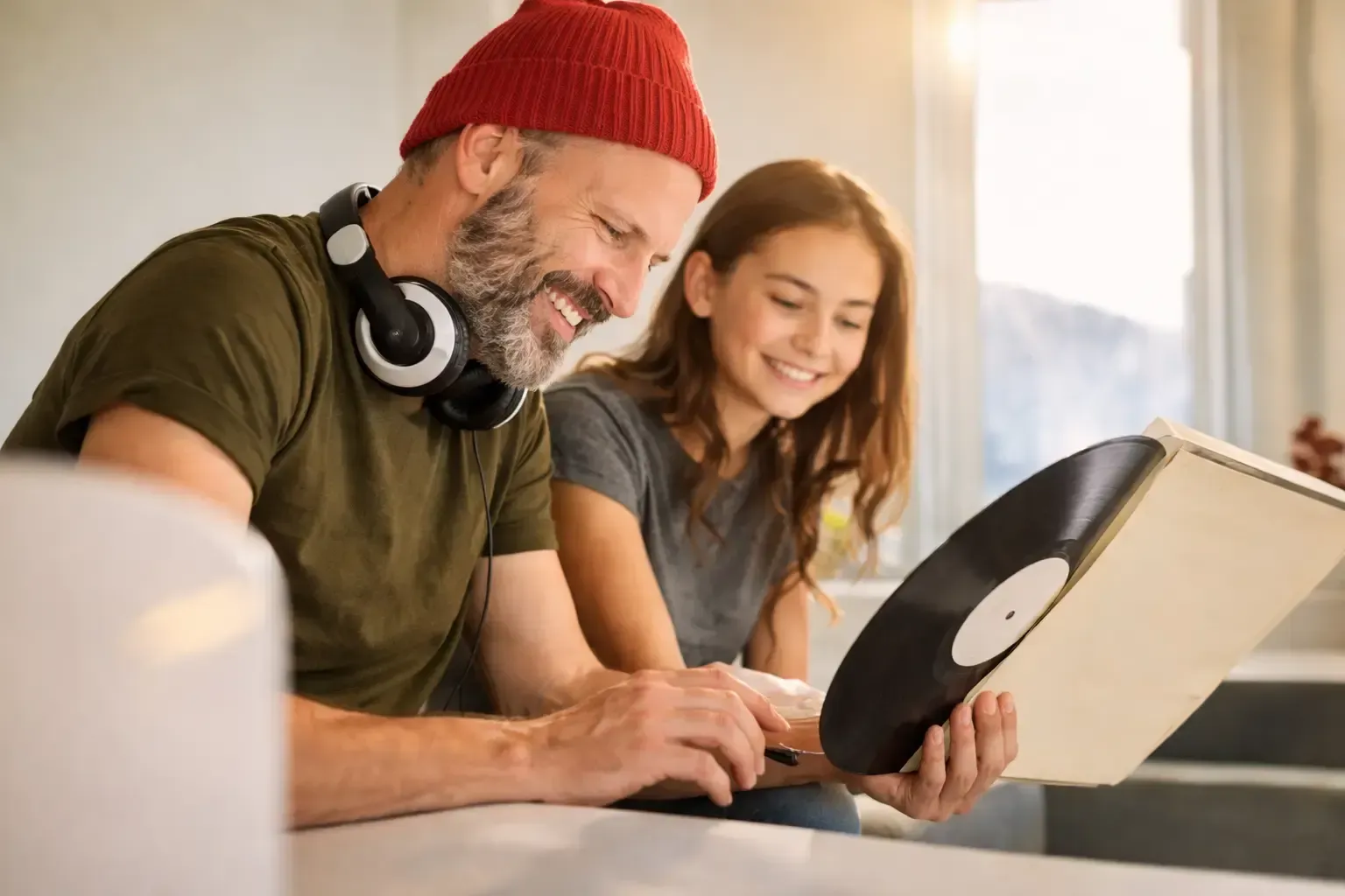 Man with beanie and headphones shows a vinyl record to a girl. Both smile, sitting indoors.