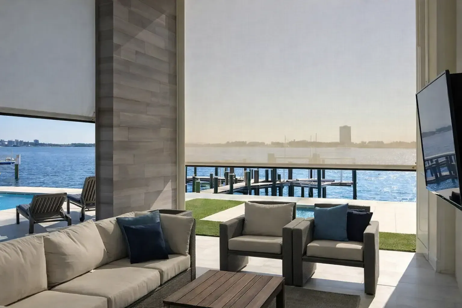 Outdoor living space with water view, beige couch, armchairs, and shades.