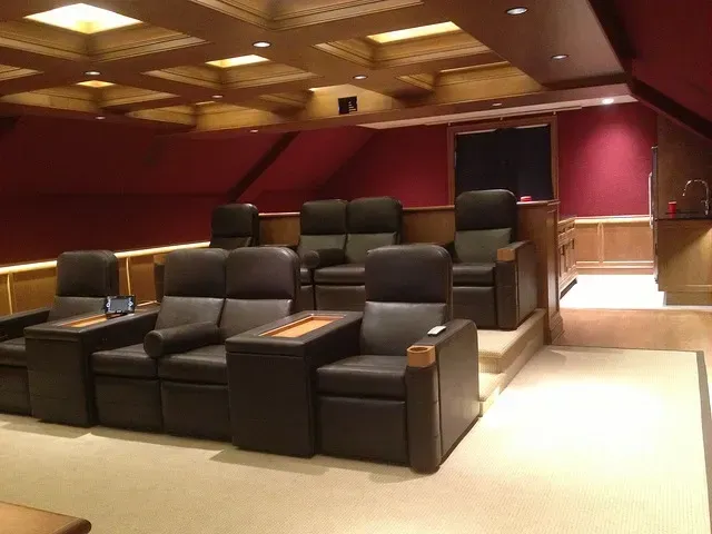 Home theater with tiered seating, black leather chairs, red walls, wooden ceiling, and entry door.