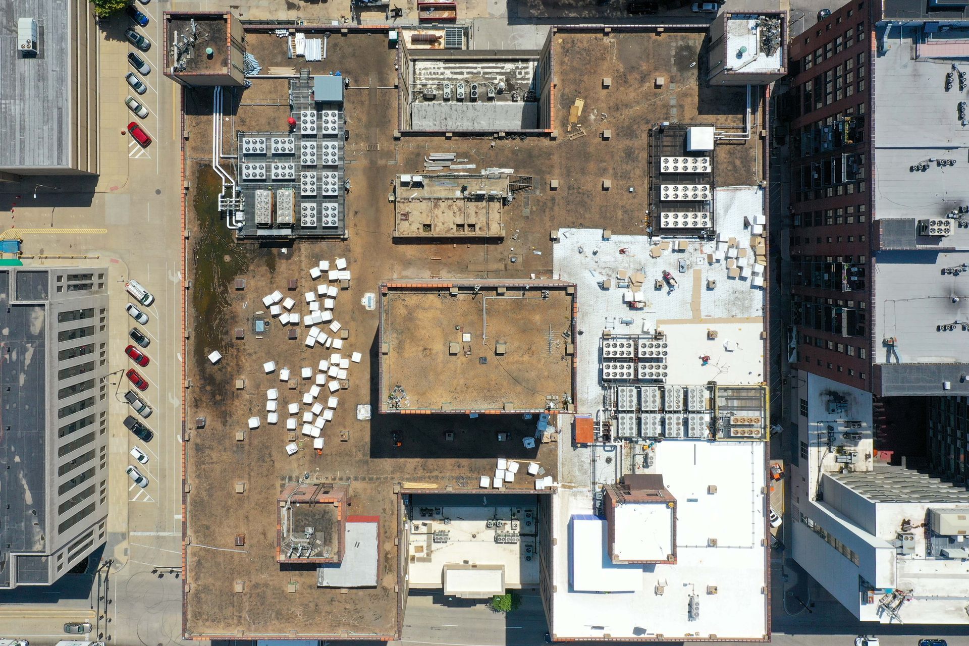 An overhead drone view of a flat building rooftop in an urban area, featuring various HVAC units and scattered debris.