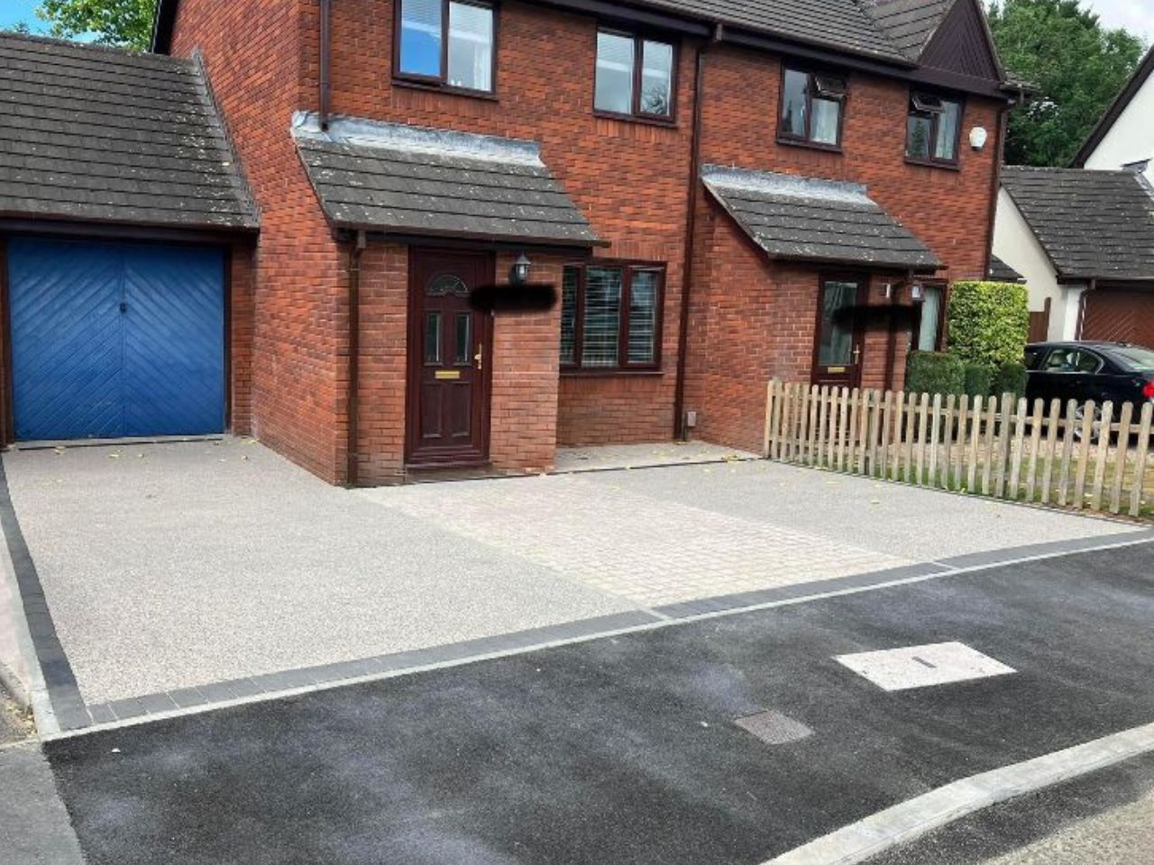 Small grey resin driveway outside of red brick house with a blue garage