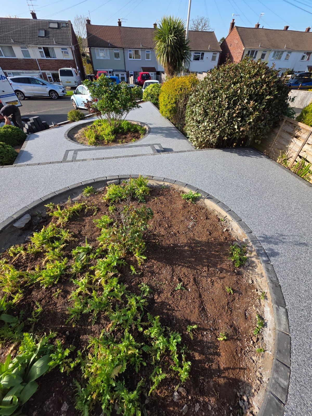 A garden area featuring two oval flower beds surrounded by grey resin paving, situated in front of residential homes.
