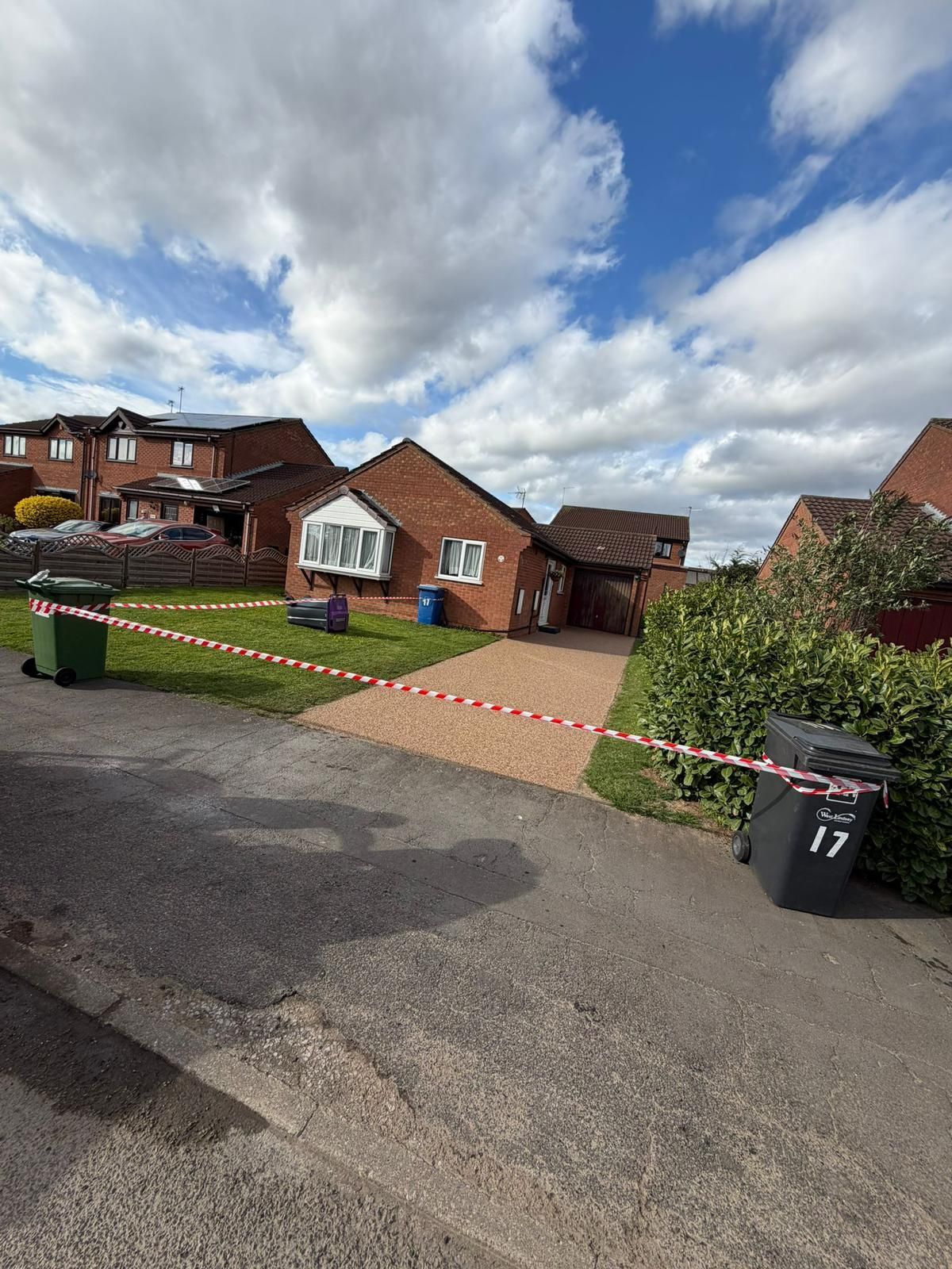 Red and white hazard tape stretches across the driveway and lawn of a brick residential house under a cloudy blue sky.