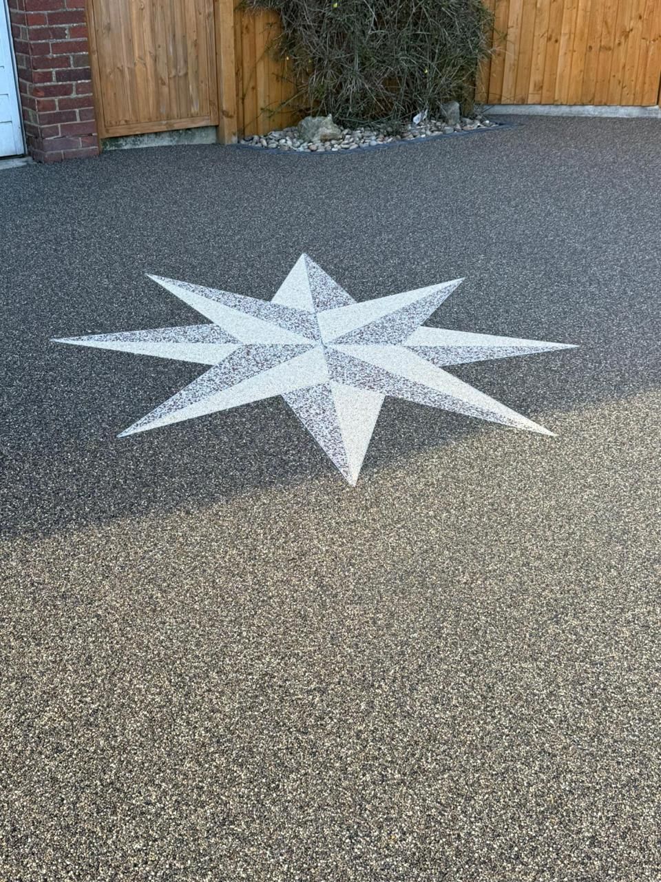 A white, eight-pointed star design embedded into a grey resin-bound driveway in front of a wooden fence and brick wall.