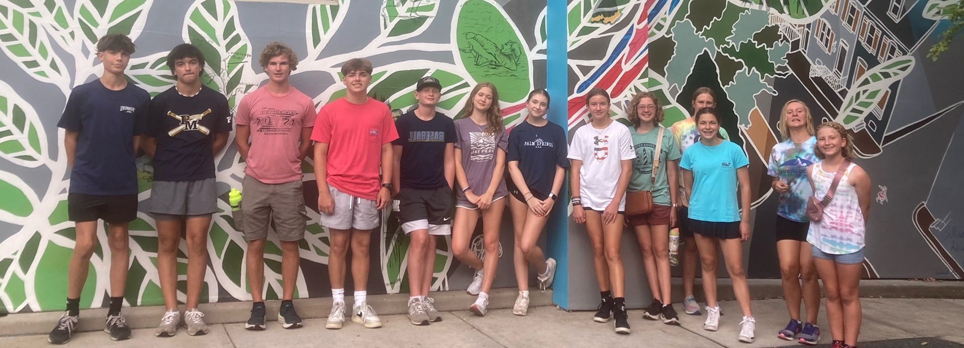 A group of teens stands in front of a mural with leaf designs and colorful abstract patterns. They wear casual clothes.