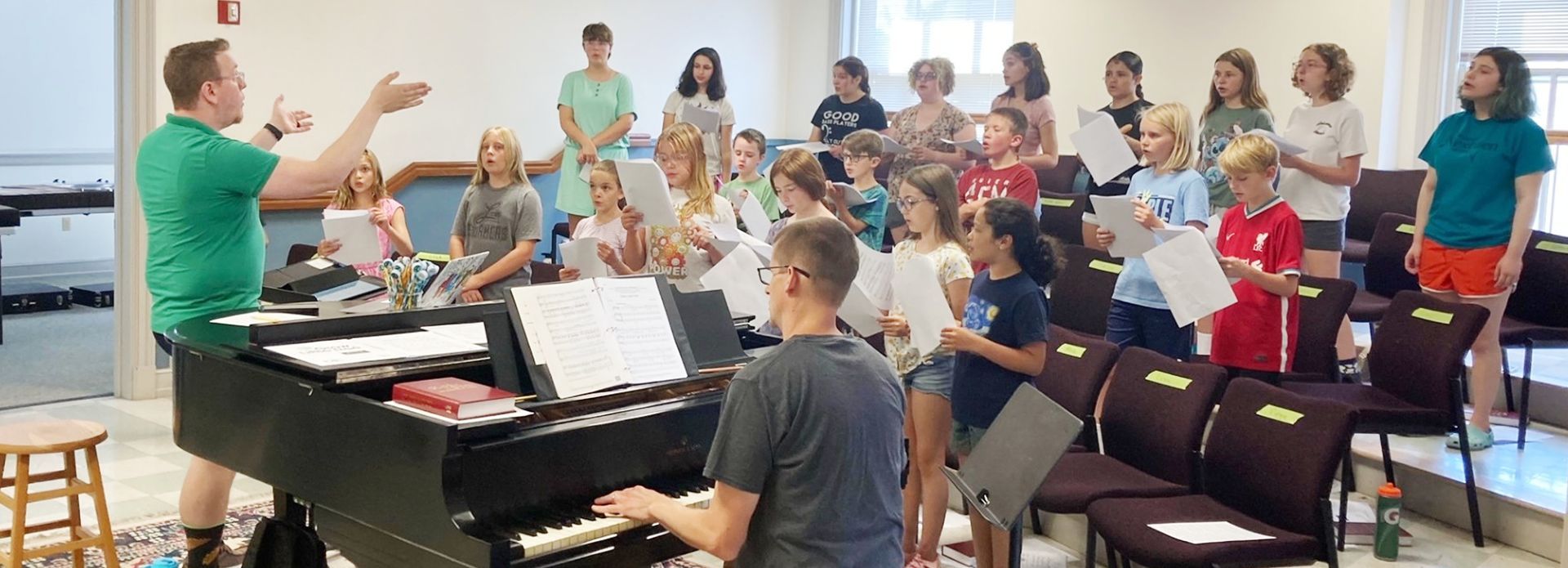 A choir of children and adults rehearsing in a room. A conductor leads while a pianist plays.