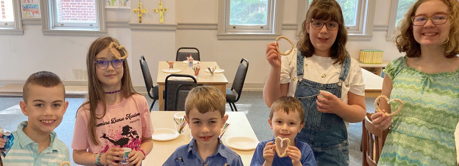 A group of children smiling at the camera. They are inside a building near tables with crafting supplies, some holding up crafts.