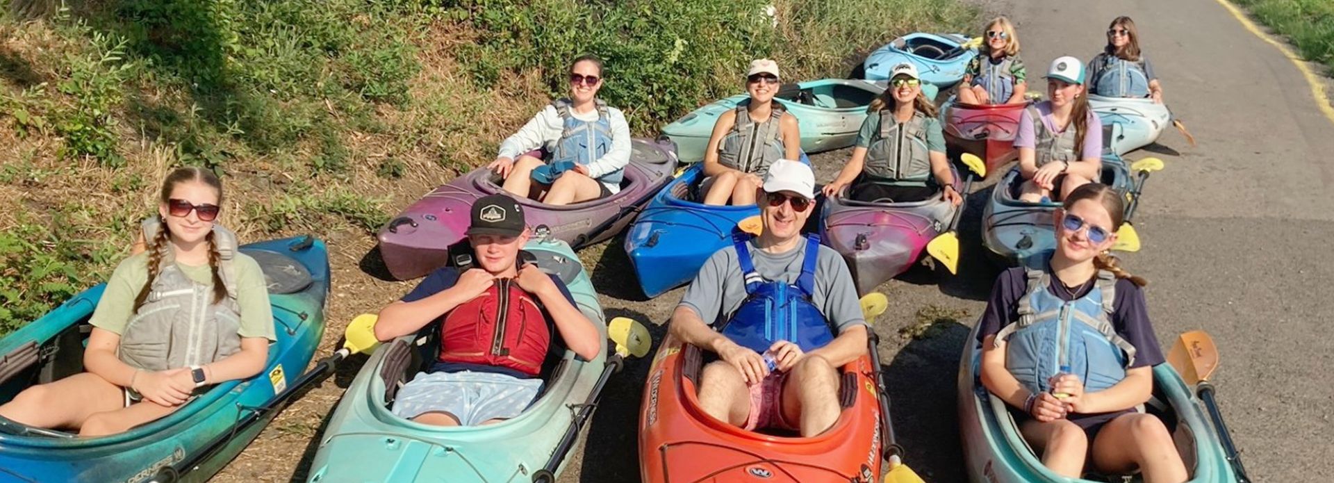 A group of people sit in kayaks on the edge of a road. They are smiling, wearing sunglasses, and dressed in casual clothing. Green foliage is visible behind them.