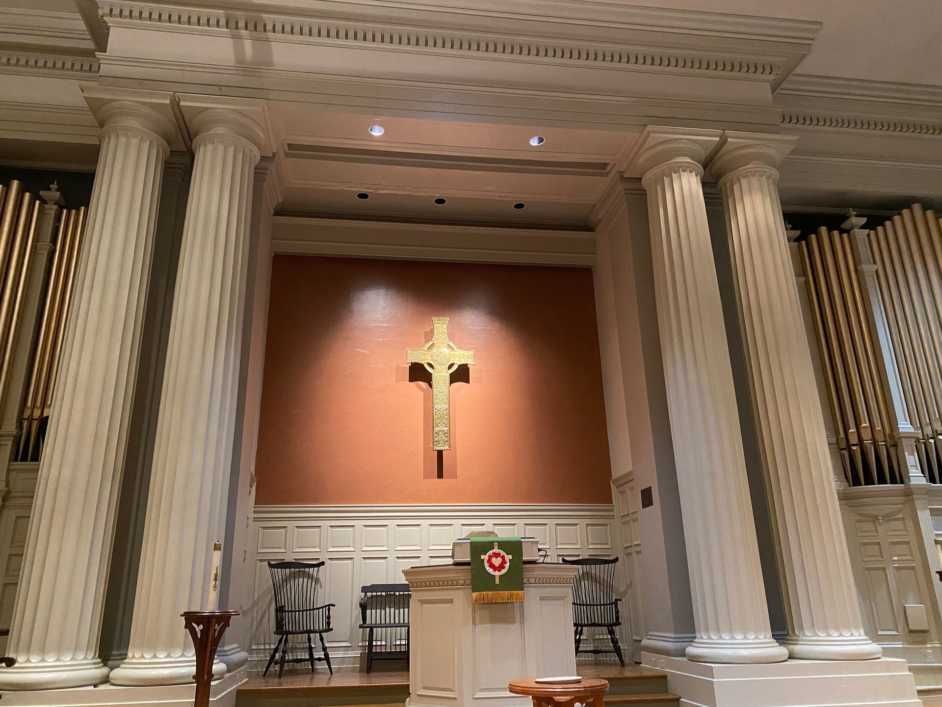 Interior of a church with white columns and a gold cross on an orange wall. A pulpit and chairs are in front of it.